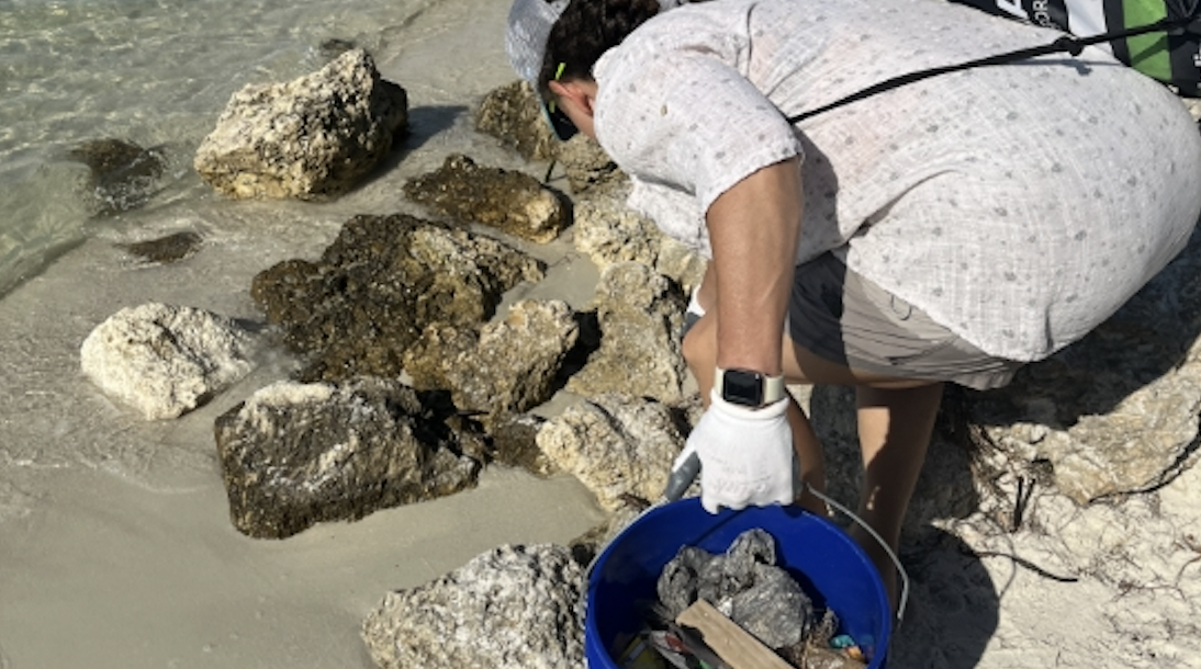 Teenage boy looks for trash and beach pollution at local Biscayne bay beach cleanup. He is holding bucket full of single-use plastic bags, bottle caps, and more.
